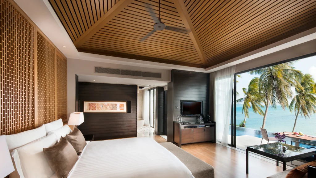 A modern hotel room with a wood-paneled ceiling, king-sized bed, and large windows opening to a balcony with palm trees and an ocean view. The room features neutral tones, a ceiling fan, and a desk area.