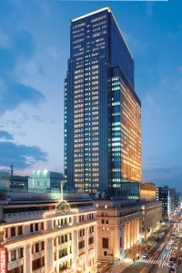 A tall, modern skyscraper with illuminated windows stands behind a historic, ornate building at dusk in a cityscape with traffic and pedestrians on the street below.