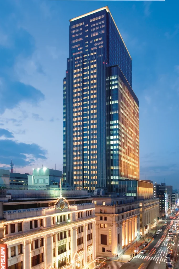 A tall, modern skyscraper with illuminated windows stands behind a historic, ornate building at dusk in a cityscape with traffic and pedestrians on the street below.