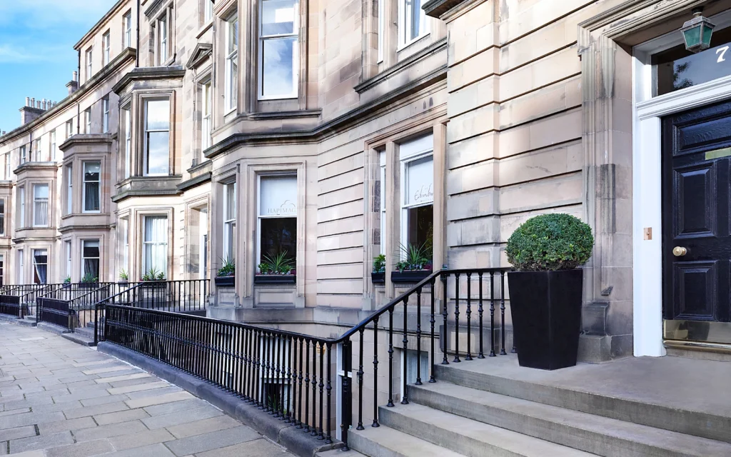 A row of elegant stone townhouses with black railings and steps, featuring large windows, potted plants, and a black door. The sidewalk runs alongside the buildings on a bright, clear day.