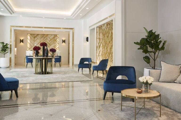 A modern, elegant hotel lobby with blue chairs, gold accents, a round table with white flowers, large plants, and a marble floor. The space is well-lit and decorated with mirrors and wall designs.