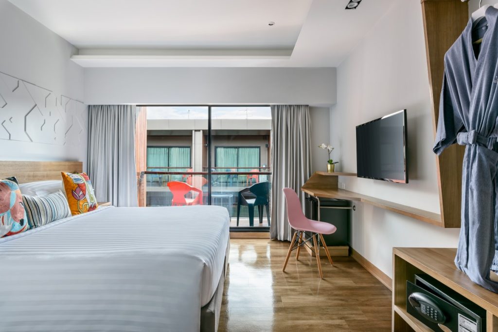 Modern hotel room with a large bed, colorful pillows, a flat-screen TV, a desk and chair, a balcony with two chairs, and a robe hanging on the wall. Light fills the space through floor-to-ceiling windows.