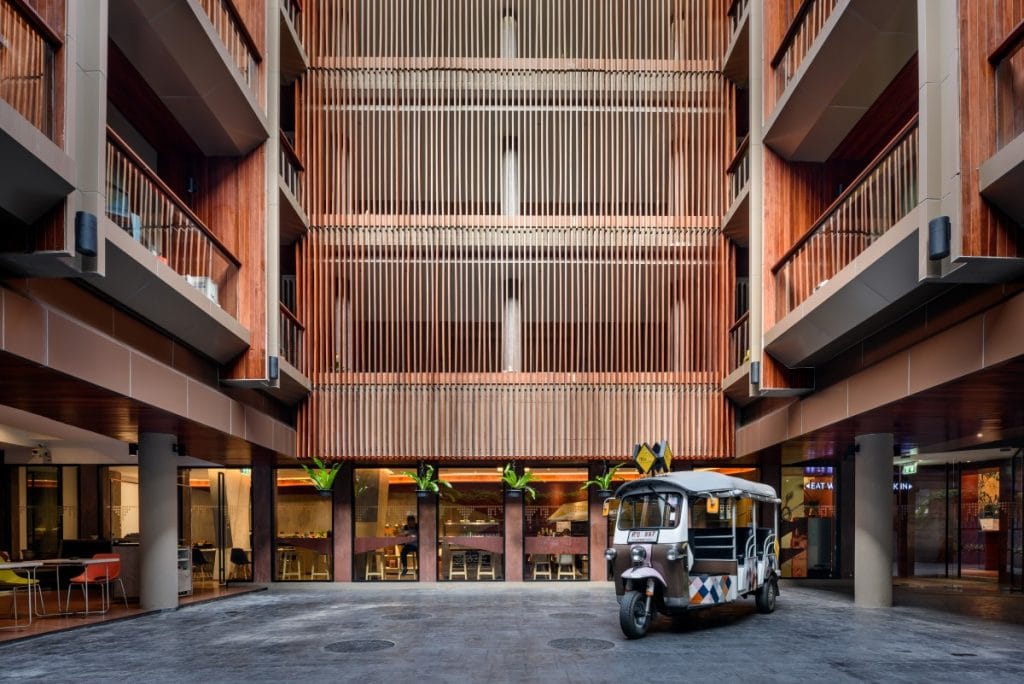 A colorful tuk-tuk is parked in the center of a modern building courtyard with wooden slats, balconies, and glass windows. The ground level has shops and seating areas with bright chairs visible.