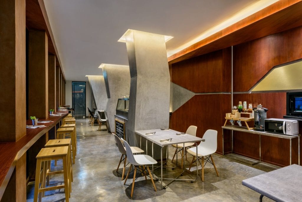 Modern dining area with wooden tables, white chairs, high stools, and unique angular concrete columns. A counter with snacks, drinks, a coffee maker, and a microwave is along the right wall. Warm lighting creates a cozy atmosphere.