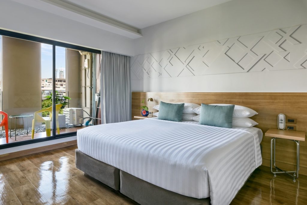 A modern hotel room with a large bed, striped white bedding, and three blue pillows, wooden flooring, a small side table, and a sliding glass door leading to a balcony with chairs and a city view.