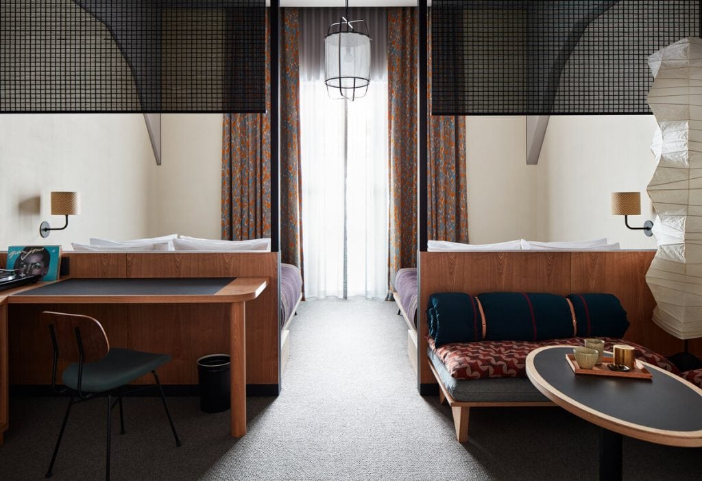 Modern hotel room with two beds separated by a partial partition, a desk with a chair on the left, a small table with tea set on the right, large window with patterned curtains, and soft lighting.