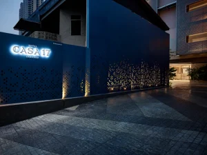 Modern exterior of Hotel Casa 17 Bangkok at dusk, featuring blue walls with a dotted pattern and illuminated signage. Warm lights shine through cut-out dots, and the entrance is visible on the right.