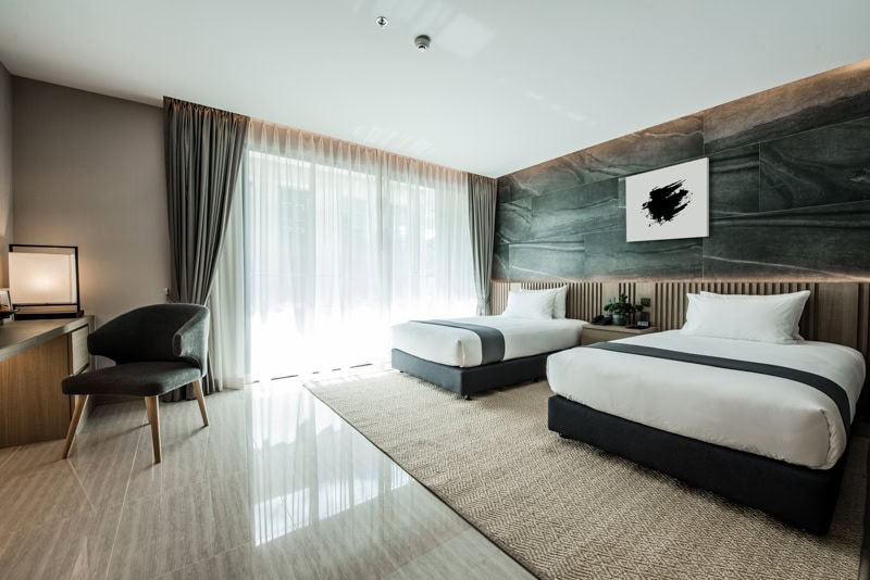 Modern hotel room with two single beds, white bedding, a desk with a chair, floor-to-ceiling window with sheer curtains, and a marble accent wall featuring abstract art above the beds.