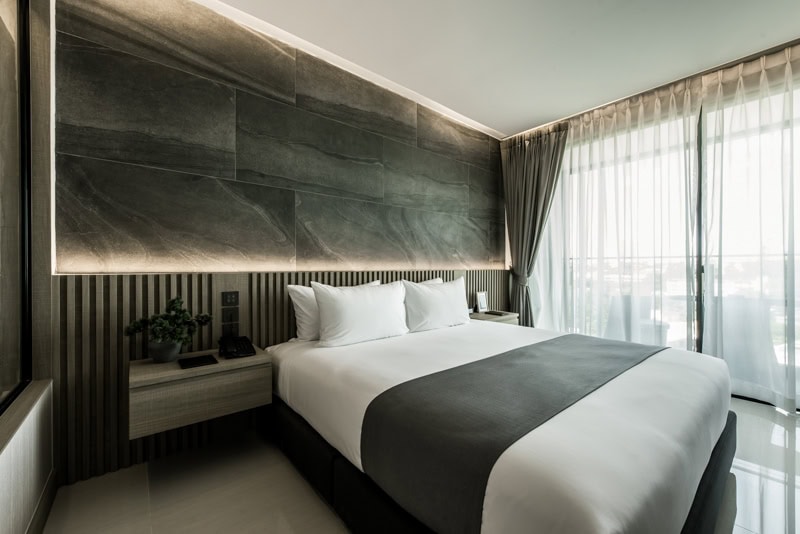 Modern hotel room with a large neatly made bed, gray and white bedding, a bedside table with a phone and plant, floor-to-ceiling windows with sheer curtains, and minimalist decor.