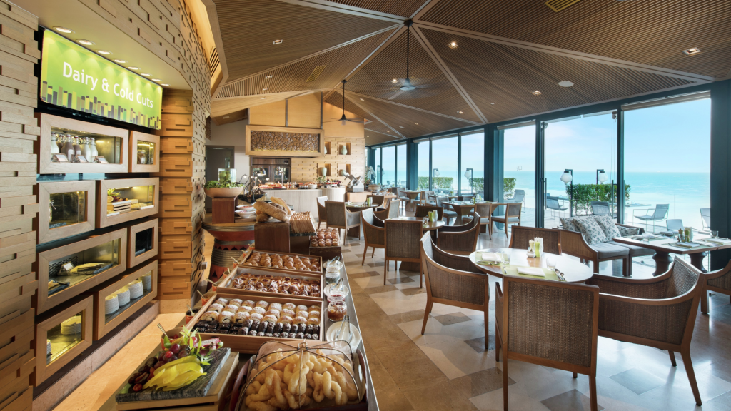 A modern, sunlit restaurant with wicker chairs and wooden tables overlooks the ocean. A buffet area features assorted pastries, dairy, and cold cuts, while large windows offer scenic sea views.