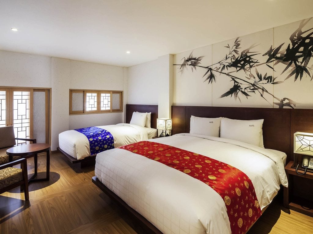A hotel room with two large beds, each with white bedding and decorative red or blue runners. The room features wooden floors, a small table with two chairs, and a wall mural of bamboo behind the beds.