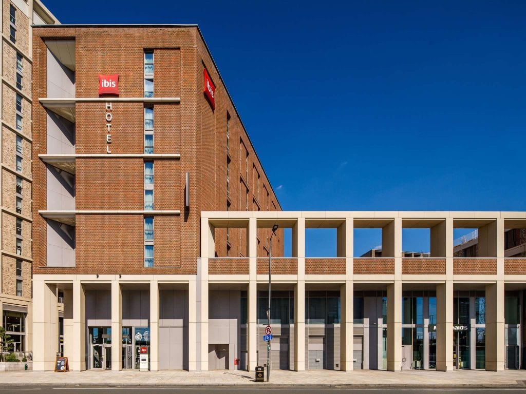 A modern brick and concrete Ibis hotel building under a clear blue sky, with large windows, a red Ibis sign, and geometric white columns at the entrance.