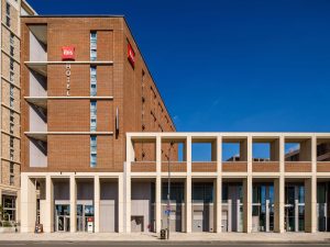 A modern brick and concrete Ibis hotel building under a clear blue sky, with large windows, a red Ibis sign, and geometric white columns at the entrance.