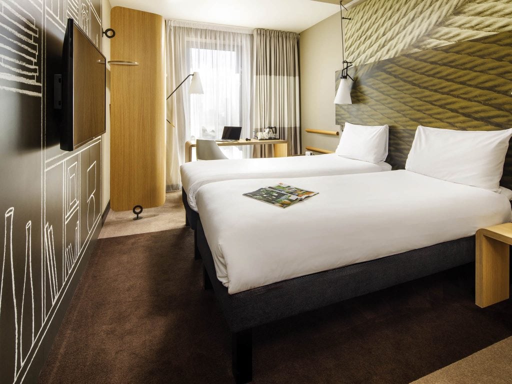Modern hotel room with two neatly made single beds, a magazine on top, a wall-mounted TV, a desk with a chair and lamp by a window with sheer curtains, and contemporary decor in neutral tones.