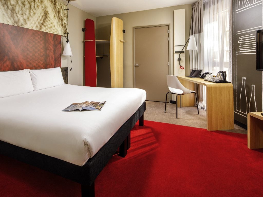 A modern hotel room with a large bed, white linens, a magazine on the bed, red carpet, desk with chair, kettle and cups, wall art, open closet, and natural light from a window with sheer curtains.
