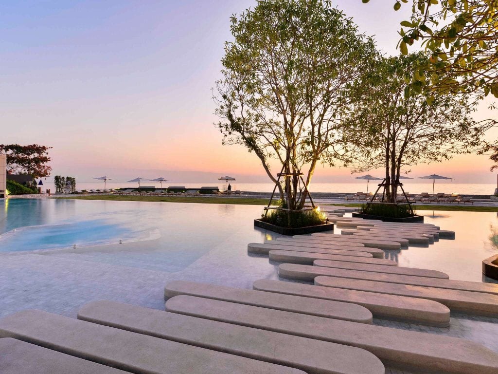 Modern pool with concrete stepping stones and small trees, overlooking a sandy beach with sun loungers and umbrellas at sunset. The sky is pastel, creating a tranquil atmosphere.