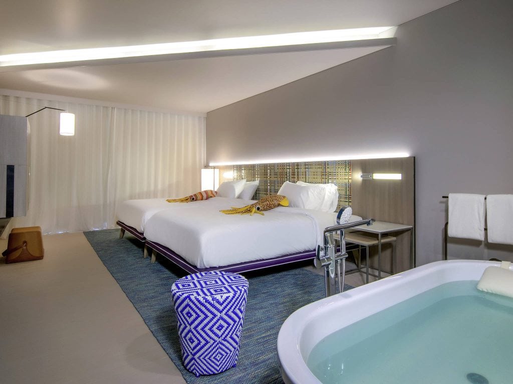 Modern hotel room with two double beds, a patterned ottoman, and a bathtub in the foreground. The room has contemporary decor, soft lighting, and a blue rug on the floor. Towels hang near the tub.
