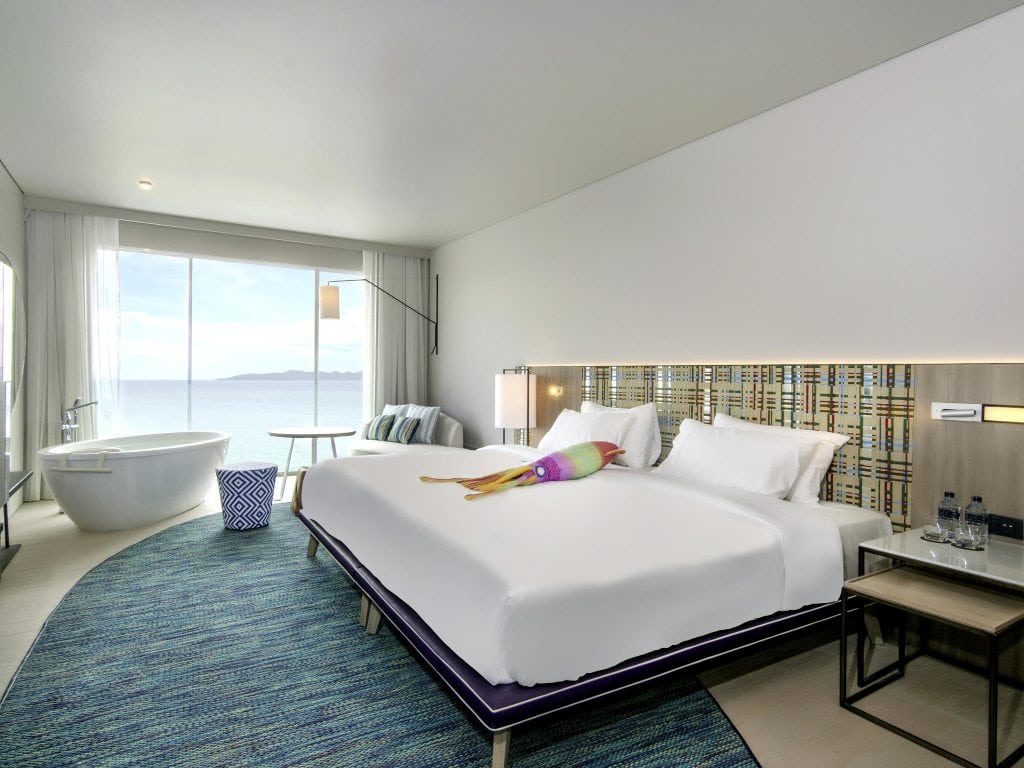 Modern hotel room with a large bed, patterned headboard, and ocean view through floor-to-ceiling windows. A freestanding bathtub sits near the window, and two bedside tables with lamps and water bottles are visible.
