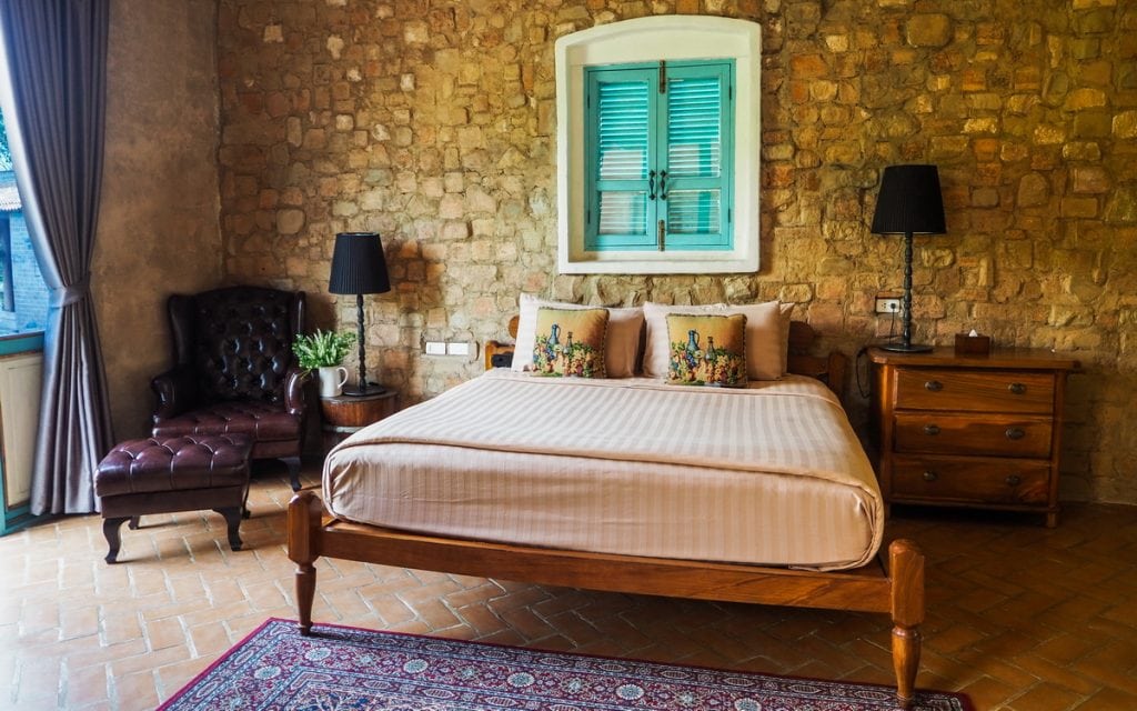 Cozy bedroom with a stone accent wall, wooden bed with striped bedding and decorative pillows, two wooden nightstands with black lamps, a leather armchair, potted plant, and a window with teal shutters.
