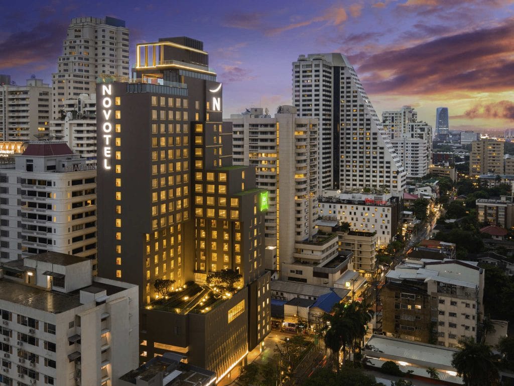 A modern cityscape at sunset features the illuminated Novotel hotel building surrounded by tall skyscrapers and city streets with cars and streetlights. The sky is a mix of dramatic clouds and vibrant colors.