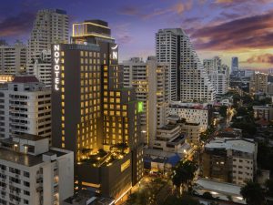 A modern cityscape at sunset features the illuminated Novotel hotel building surrounded by tall skyscrapers and city streets with cars and streetlights. The sky is a mix of dramatic clouds and vibrant colors.
