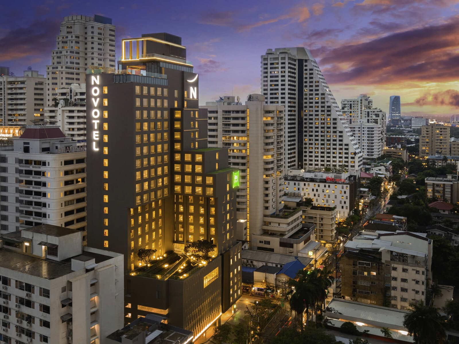 A modern cityscape at sunset features the illuminated Novotel hotel building surrounded by tall skyscrapers and city streets with cars and streetlights. The sky is a mix of dramatic clouds and vibrant colors.