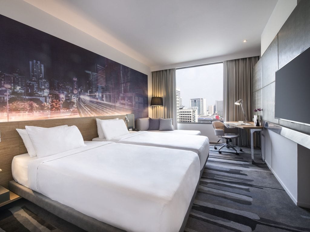Modern hotel room with two neatly made double beds, a cityscape mural on the wall, a large window with a city view, a desk and chair, flat-screen TV, and contemporary decor in neutral tones.