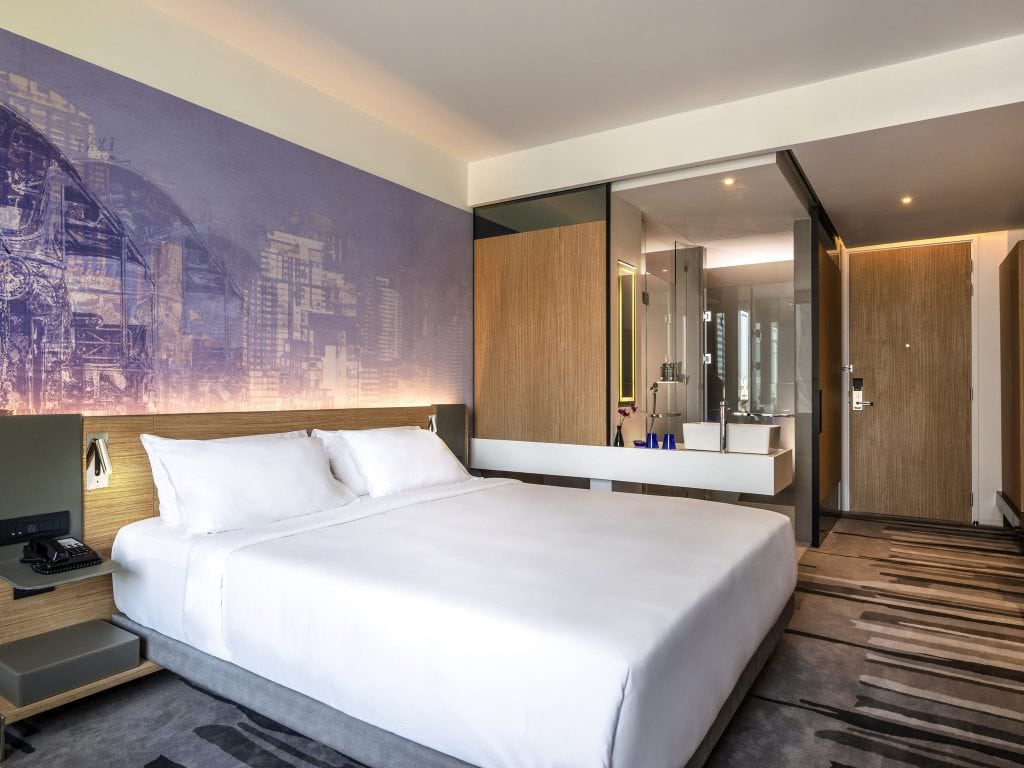 Modern hotel room with a large bed, white bedding, bedside tables, a lighted purple wall mural, and an open bathroom area with glass walls and a vanity sink. The room has a minimal, contemporary design.