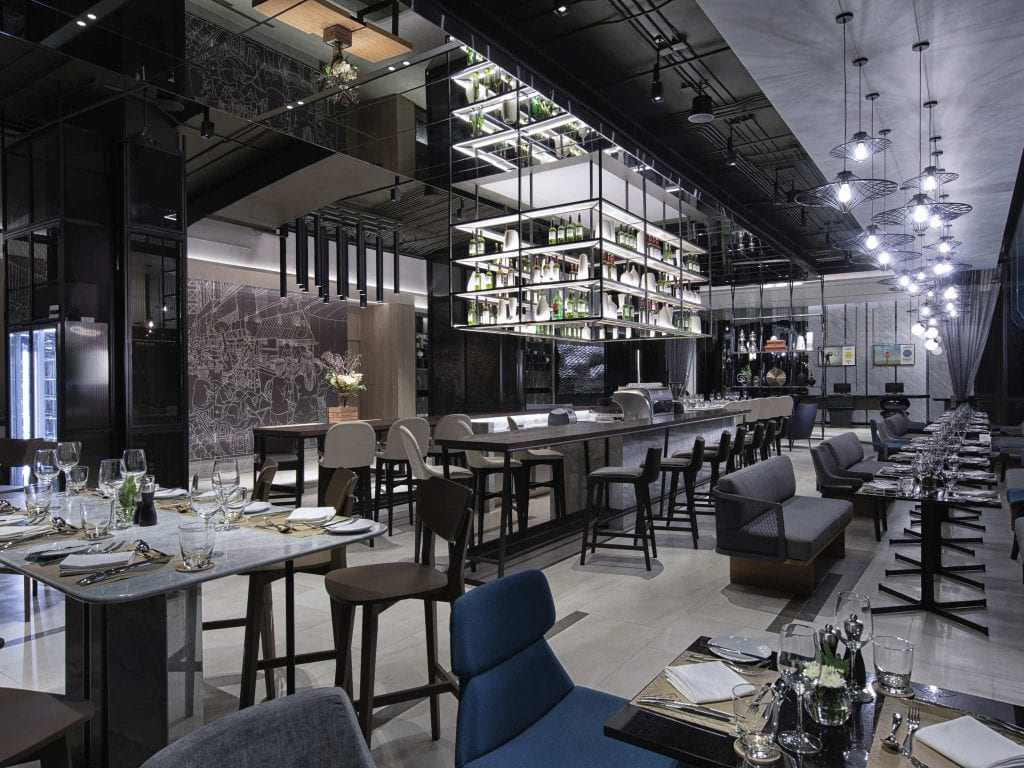 A modern, upscale restaurant interior with a central bar, high shelves of bottles, elegant tables set for dining, stylish lighting, and contemporary decor in dark and neutral tones.