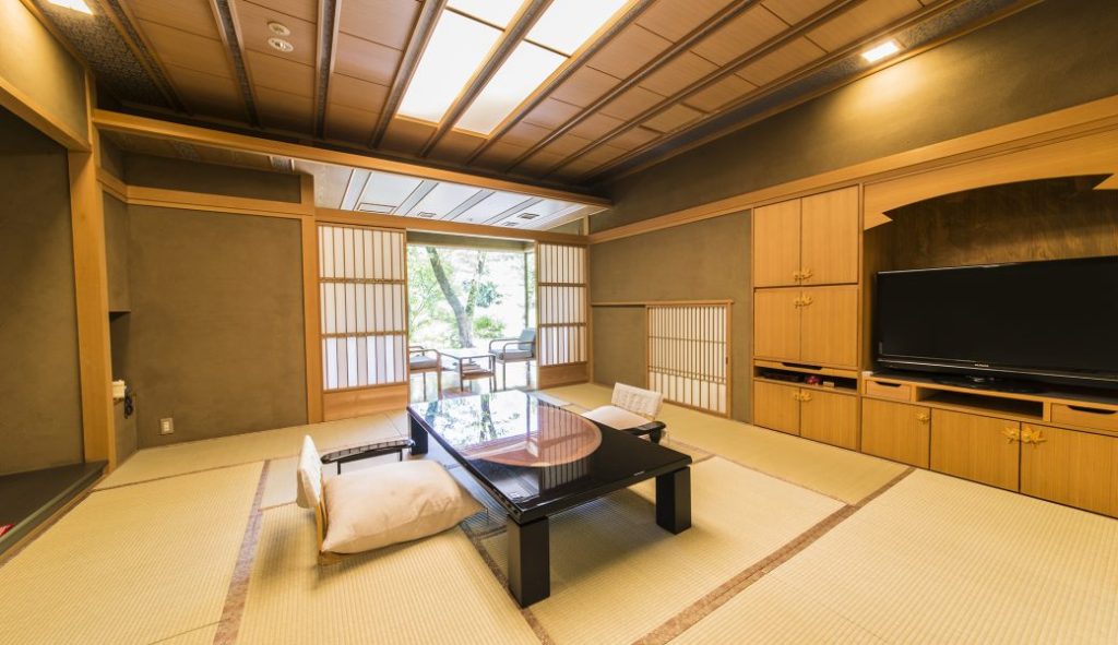 A traditional Japanese tatami room with a low black table, floor cushions, sliding shoji doors, built-in wooden cabinets, and a TV. Natural light fills the room, and there are chairs near the window overlooking a garden.