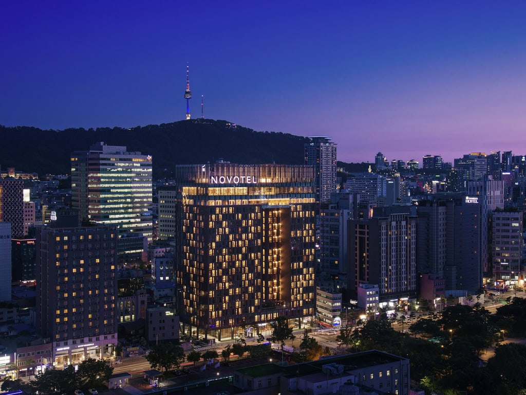 A cityscape at dusk in Seoul, featuring the illuminated Novotel hotel in the foreground, surrounded by tall buildings, with Namsan Seoul Tower visible on a hill in the background.