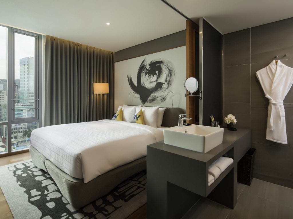A modern hotel room with a large bed, abstract artwork above the headboard, a tall window with city views, and a sleek sink area with a white robe hanging on the wall.