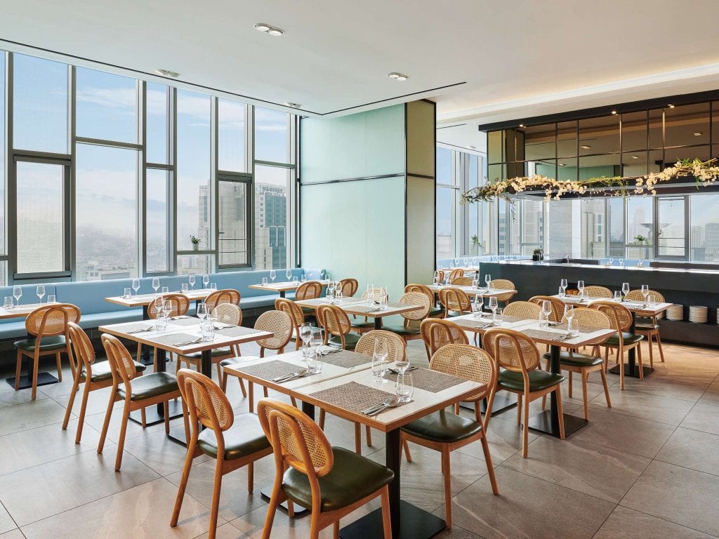 Modern restaurant with large windows offering a city view, wooden tables and chairs neatly arranged, wine glasses set on the tables, and a stylish bar area decorated with hanging greenery.
