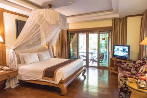 A cozy hotel room with a large canopy bed, wooden floors, bedside tables, a sofa with floral upholstery, a TV, lamps, and glass doors opening to a balcony with outdoor seating and greenery outside.