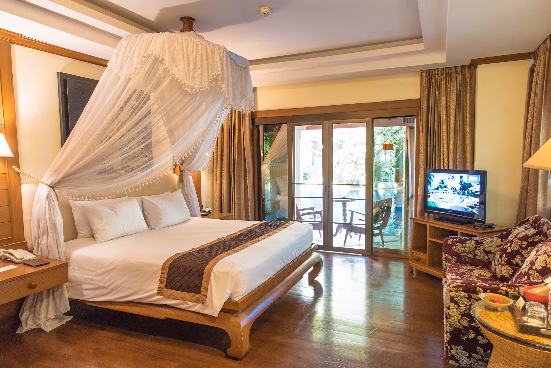 A cozy hotel room with a large canopy bed, wooden floors, bedside tables, a sofa with floral upholstery, a TV, lamps, and glass doors opening to a balcony with outdoor seating and greenery outside.