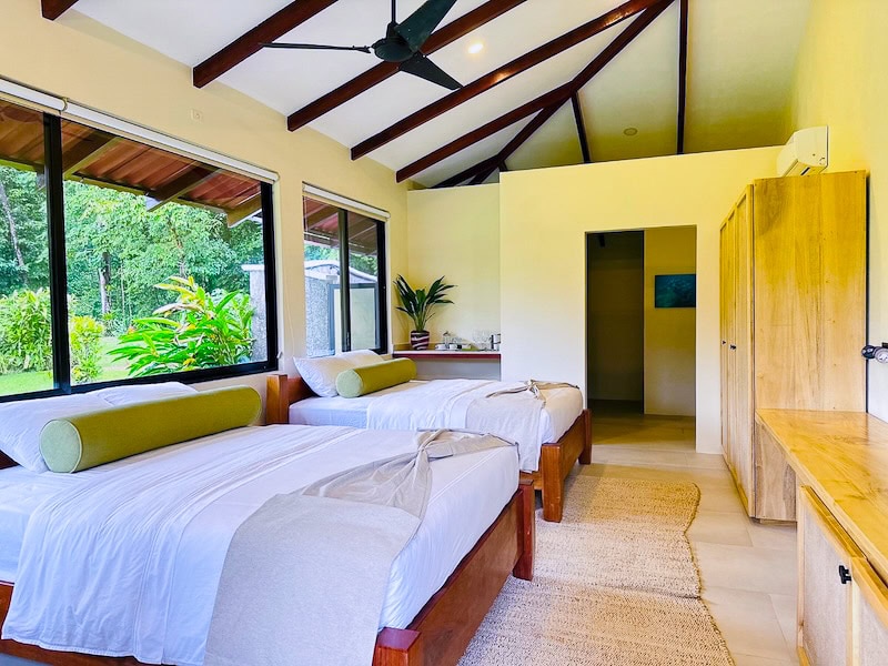 A bright bedroom with two wooden beds, green bolster pillows, a woven rug, large windows showing lush greenery, wooden wardrobe, and vaulted ceiling with exposed beams and a ceiling fan.