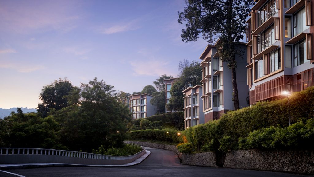 A winding road curves uphill past modern multi-story buildings with large windows, surrounded by lush greenery and tall trees, under a clear evening sky.