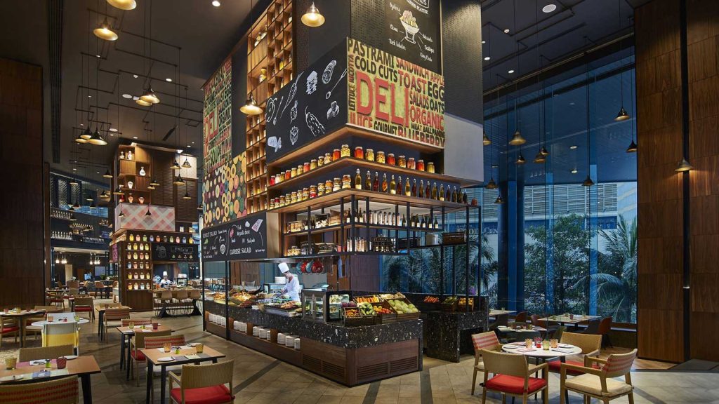 Modern restaurant interior with high ceilings, large windows, and stylish lighting. An open kitchen and deli counter are surrounded by neatly arranged tables and chairs. Colorful wall art and shelves of jars add a lively touch.