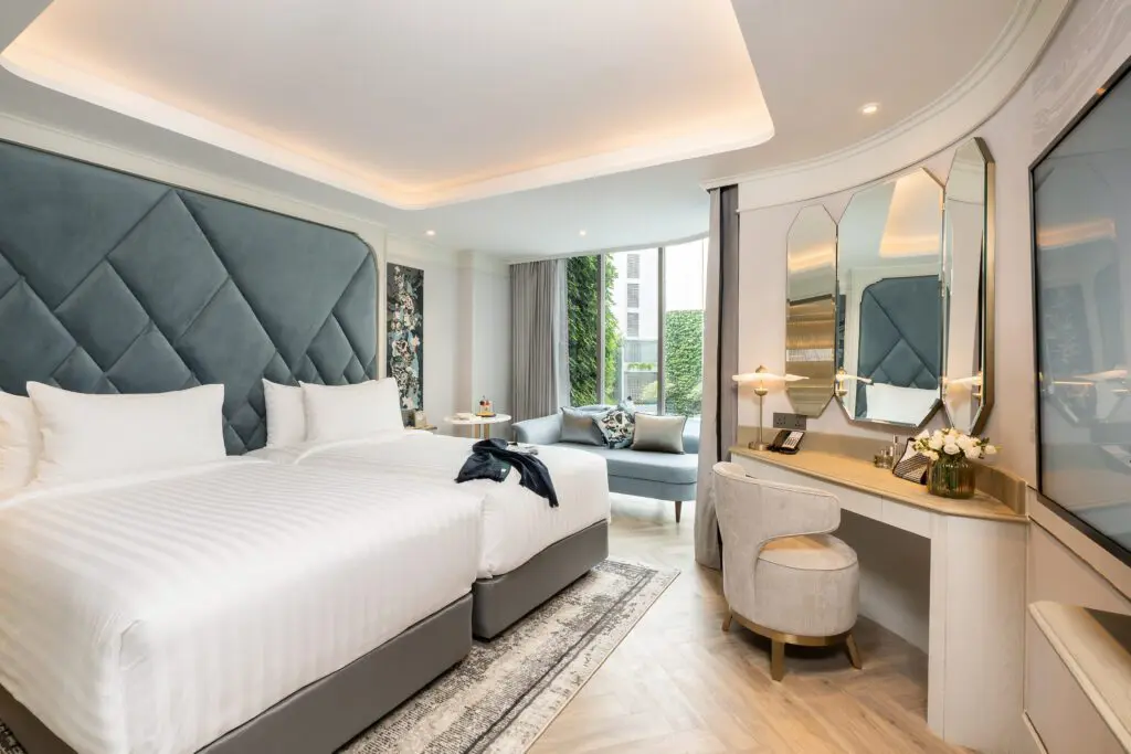 A modern hotel room with two large beds, a stylish headboard, a lounge chair by a window, a vanity desk with a chair, mirrors, and fresh flowers; the room is bright and elegant with neutral colors.