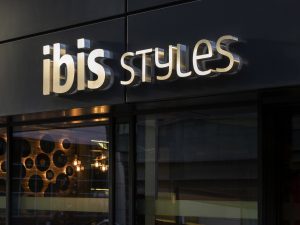 The illuminated sign of ibis Styles hotel is mounted on a dark exterior wall above large glass windows, with reflections of lights and modern decor visible inside.