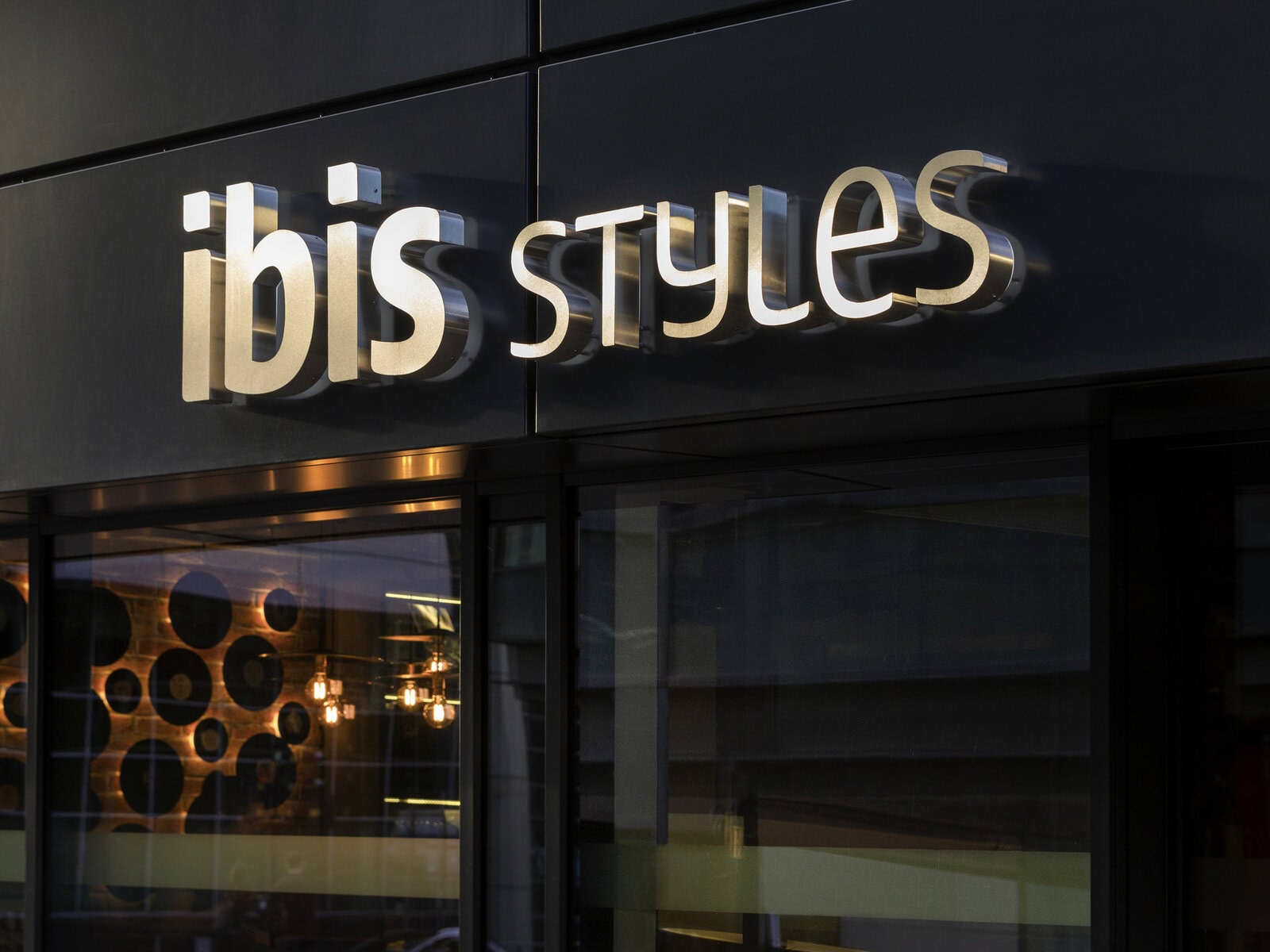 The illuminated sign of ibis Styles hotel is mounted on a dark exterior wall above large glass windows, with reflections of lights and modern decor visible inside.