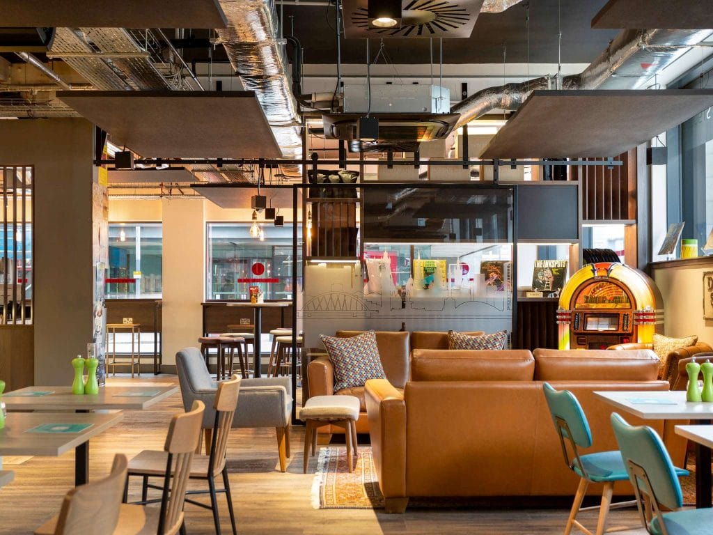 Modern lounge with tan leather sofas, blue chairs, and wooden tables. A vintage jukebox sits in the corner, and magazines are displayed on a glass wall. The space is bright with industrial-style ceiling fixtures.