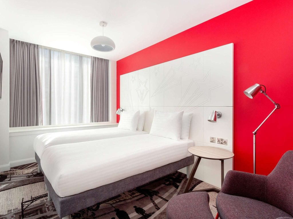 A modern hotel room with two single beds pushed together, white bedding, a bright red accent wall, white headboard, gray curtains over a large window, a round bedside table, and a gray armchair with a floor lamp.