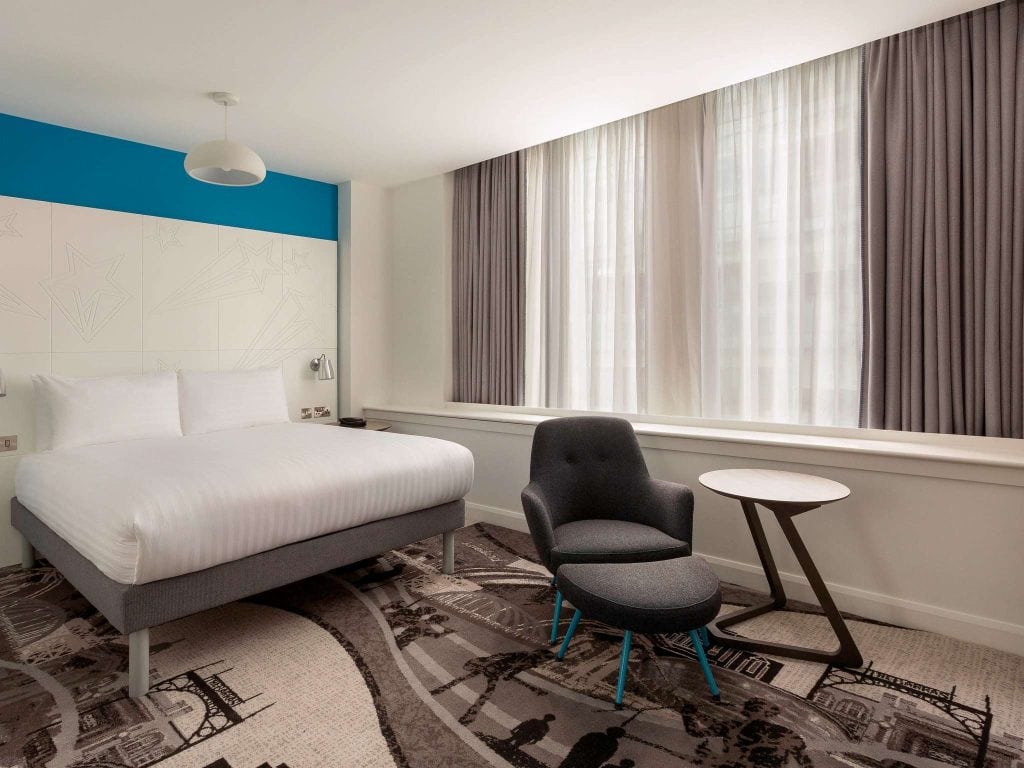 Modern hotel room with a double bed, white bedding, a gray armchair with ottoman, a small round table, and large windows with white sheers and gray curtains. The carpet has a cityscape pattern.