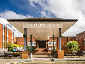 Entrance of a Mercure Hotel with a covered walkway, outdoor seating, plants, and brick building under a partly cloudy sky.