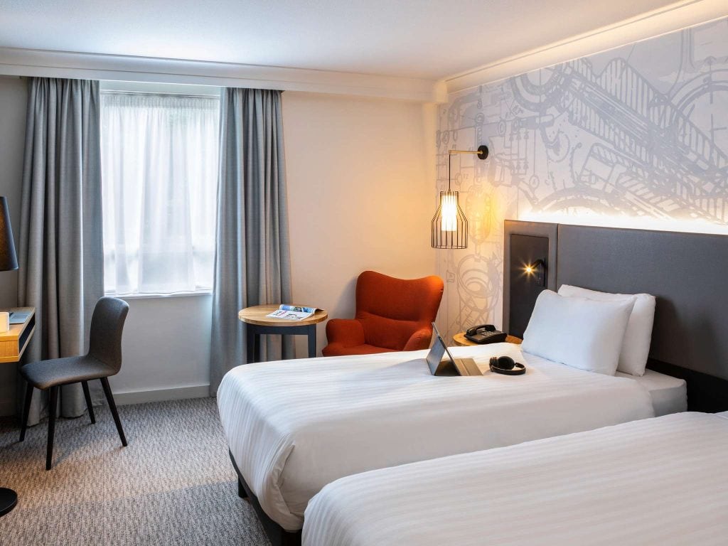 A modern hotel room with two single beds, a red armchair by a small round table, a desk with a chair, a window with gray curtains, and soft lighting. A laptop and headphones are on one bed.