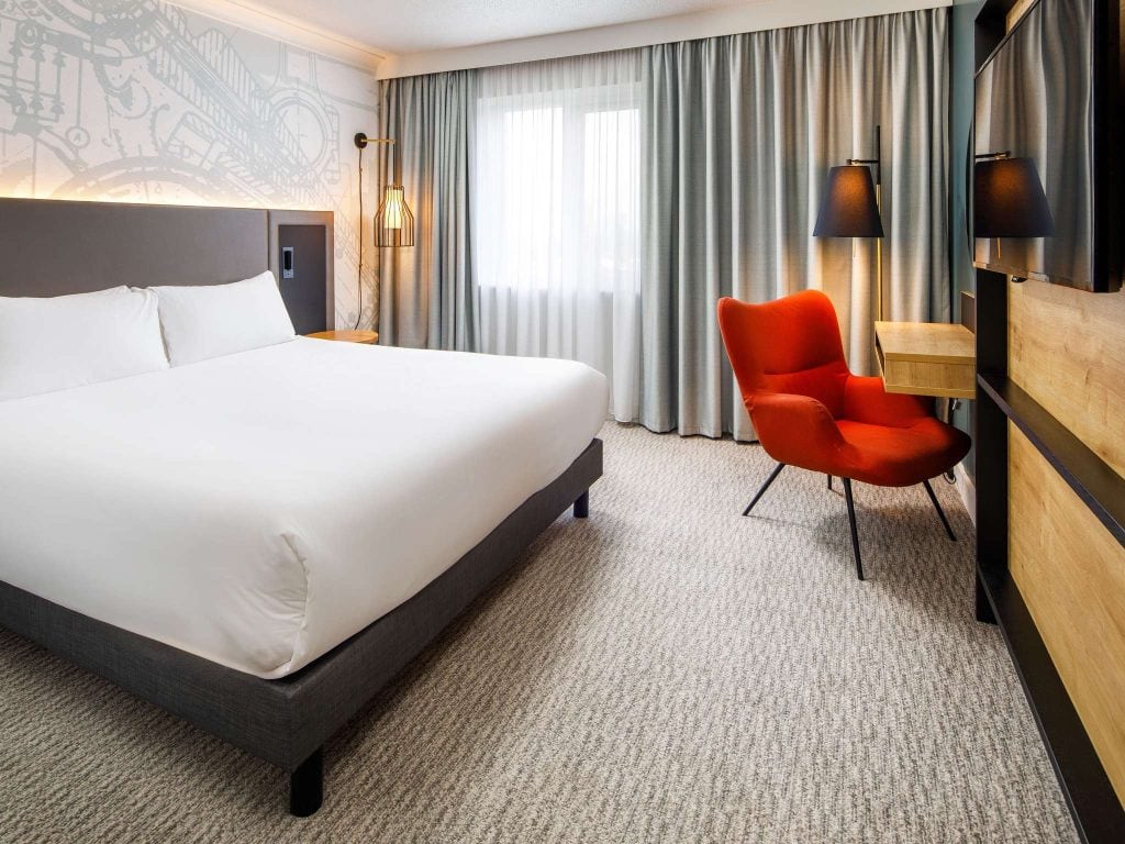 A modern hotel room with a large bed, gray carpet, a wooden desk, an orange chair, two black lamps, and a window with light gray curtains. The wall behind the bed has a subtle clock-themed design.