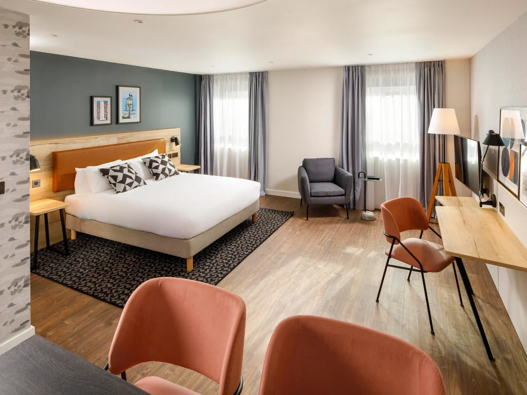 Modern hotel room with a large bed, two patterned pillows, gray armchair, tall lamp, long desk, TV, and pink chairs. The room has wood flooring, large windows with gray curtains, and minimalist decor.