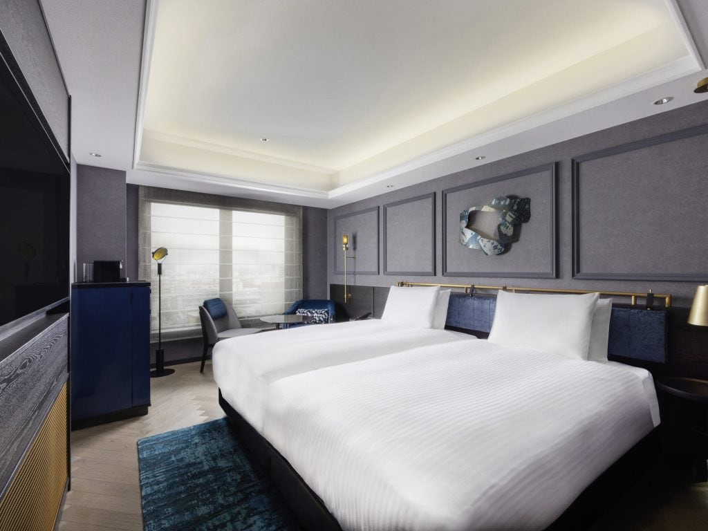 A modern hotel room with a large bed, navy and gray decor, a blue cabinet, a window with sheer curtains, a unique wall mirror, and a sitting area with a chair and lamp.