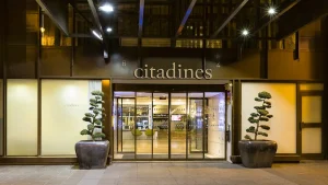 The entrance of a Citadines hotel at night, featuring glass doors, large windows, two potted topiary plants, and warm lighting inside. The hotel name citadines is displayed above the entrance.